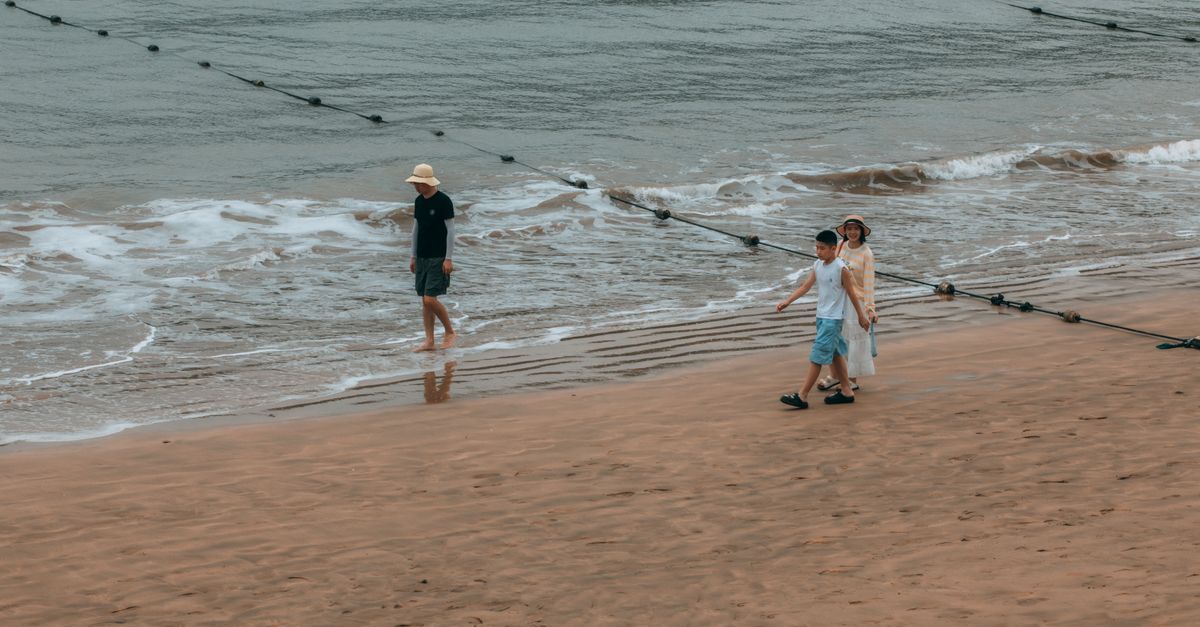 Three people in sun hats walk through the receding waves on the wet sand of a wide beach