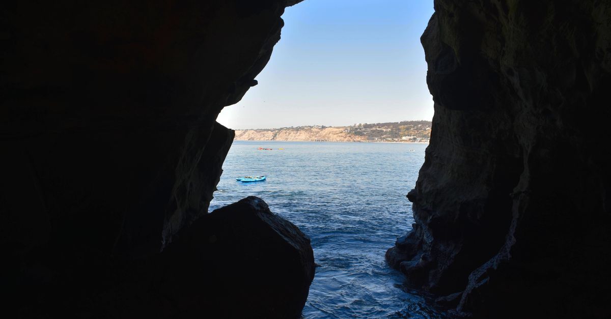 Two kayaks gliding through a narrow rock arch, with an excursion boat waiting off the ochre cliffs behind them
