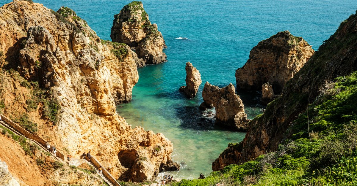 Stone steps winding down a green cliffside towards a turquoise cove ringed by rock pillars