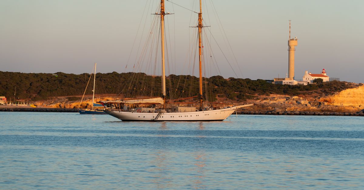 A sailing boat with its mainsail set cruises along a rocky Portuguese cliff coast in the afternoon light